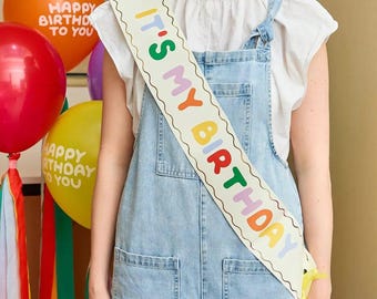 Può includere: Una fascia di compleanno bianca con il testo IT'S MY BIRTHDAY in lettere colorate e arrotondate. La fascia presenta un bordo ondulato con contorno scuro ed è indossata sopra un top bianco a maniche corte e una salopette in denim azzurro.