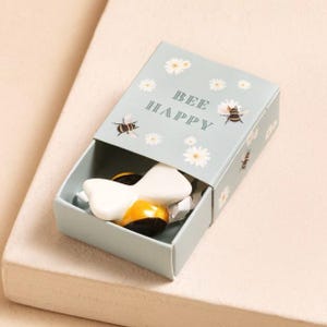 May include: A light blue matchbox-style gift box with the words "BEE HAPPY" and illustrations of bees and daisies. Inside the box is a white and yellow bee-shaped object.