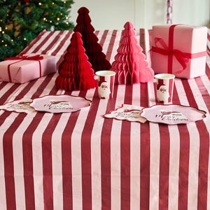 May include: A table setting featuring a red and white striped tablecloth, two paper cups with Santa Claus illustrations, two scalloped plates with Merry Christmas text, two honeycomb paper trees in red and pink, and two pink gift boxes with red ribbons.