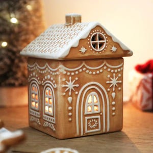 Può includere: Una casetta di pan di zenzero in ceramica con base marrone chiaro e dettagli in glassa bianca. La casa presenta un tetto innevato, finestre e un camino. Le finestre si illuminano di una luce calda, creando una decorazione festosa.