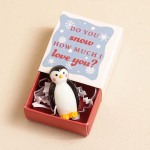 Puede incluir: Una figura de pingüino de cerámica dentro de una caja de cerillas roja. El pingüino tiene un cuerpo blanco, alas negras y patas amarillas. La tapa de la caja de cerillas tiene el texto "Do you snow how much I love you?" en rojo y azul.