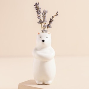 May include: A white ceramic polar bear vase holding dried lavender stems. The vase has a minimalist design with a black nose and eyes. The bear's arms are crossed over its chest. The vase is on a light beige surface.
