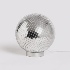 May include: A spherical disco ball lamp featuring numerous small, reflective silver mirror tiles covering the entire surface. The round light fixture is mounted on a circular, metallic silver base and includes a thin white power cord extending outward.