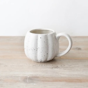 May include: A white ceramic mug shaped like a pumpkin, with a handle. The mug features a speckled finish with small black dots. It rests on a light-coloured wooden surface.
