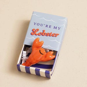 May include: A novelty gift set featuring a bright orange lobster figurine inside a blue matchbox. The matchbox has the text "YOU'RE MY Lobster" in orange script. The box has a blue and white striped design.