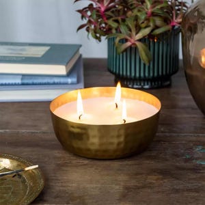May include: A gold metal candle holder with three lit candles inside. The candle holder is on a wooden surface.