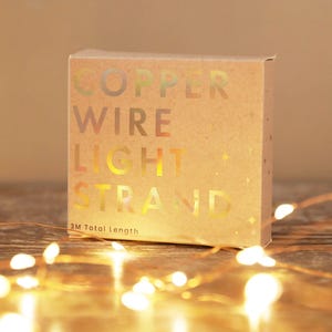 Op de afbeelding: Een kartonnen doos met de woorden "COPPER WIRE LIGHT STRAND" in goud gedrukt. De doos staat op een houten oppervlak met een streng kleine, warmwitte lampjes. De tekst "3M Total Length" staat onderaan.