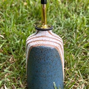 May include: A ceramic oil bottle with a blue base and a white and brown striped upper section. It has a gold spout with a black tip, designed for dispensing oil or vinegar. The bottle is set against a backdrop of green grass.