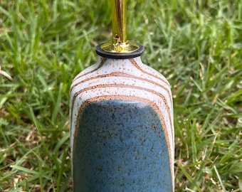 Può includere: Un'oliera in ceramica con una sezione inferiore blu maculata e una sezione superiore bianca con strisce orizzontali marroni. La bottiglia è sormontata da un beccuccio versatore in metallo dorato con un piccolo tappo nero sulla punta.