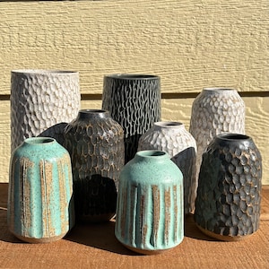 May include: A collection of ten ceramic vases in various colors and textures. The vases are arranged in a row on a wooden surface. The vases are all different shapes and sizes, and some have a textured surface.