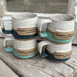 May include: Four white ceramic mugs with speckled glaze. Each mug has a band of turquoise and dark blue glaze and a brown label with the text "Memphis Magnetic Recording Co."