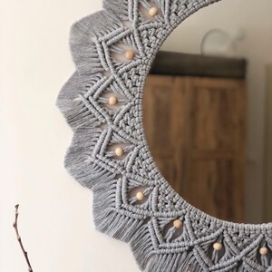 May include: A gray macrame mirror with a circular frame and wooden beads. The mirror has a fringe border and a geometric pattern.