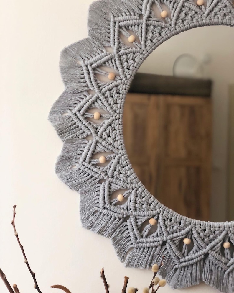 May include: A gray macrame mirror with a circular frame and wooden beads. The mirror has a fringe border and a geometric pattern.