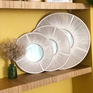 May include: Three white rope circular mirrors of different sizes are displayed on a wooden shelf against a yellow wall. The mirrors are arranged in a staggered pattern, with the largest mirror at the back and the smallest mirror in the front.
