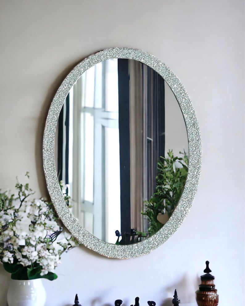 Puede incluir: Un espejo ovalado con un marco de purpurina plateada. El espejo est&aacute; colgado en una pared blanca y refleja una ventana con vista a la vegetaci&oacute;n.