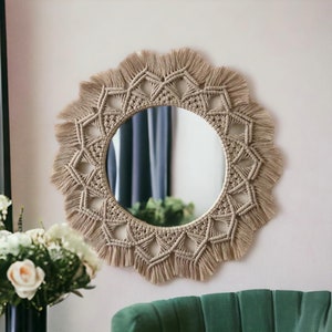 May include: A round macrame mirror with a natural beige colour and a fringe border. The mirror is hanging on a white wall.