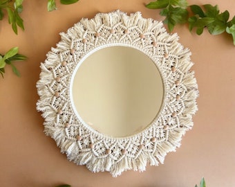 May include: A round mirror with a white macrame frame and wooden beads. The mirror has a white background and is surrounded by a fringe.