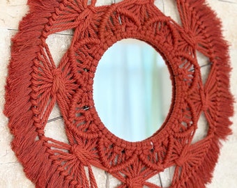 Puede incluir: Un espejo redondo con un marco de macramé de color naranja quemado. El marco tiene un patrón geométrico y un borde de flecos.