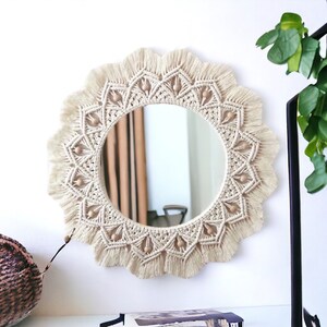 May include: A round macrame mirror with a natural beige color and wooden beads around the edge. The mirror has a fringe border.