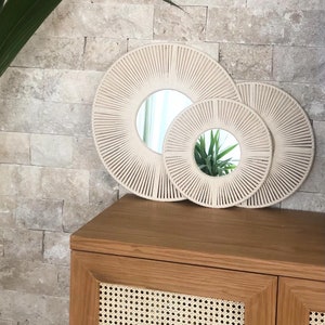 May include: Two round mirrors with a woven rope frame. The larger mirror is beige and the smaller mirror is white. Both mirrors are hanging on a beige tiled wall.