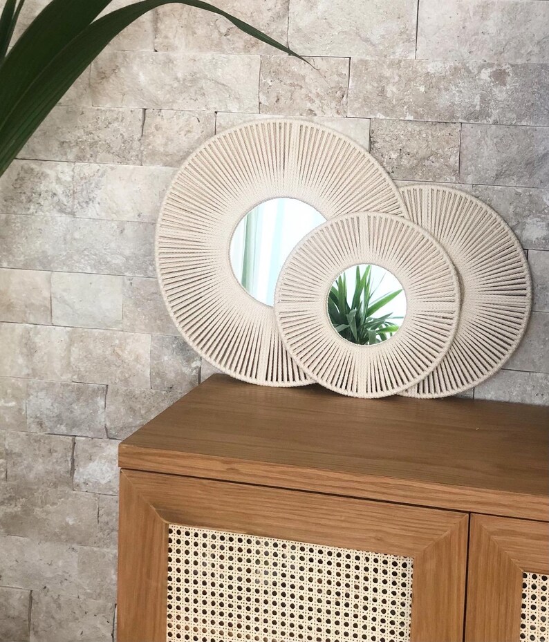 May include: Two round mirrors with a woven rope frame. The larger mirror is beige and the smaller mirror is white. Both mirrors are hanging on a beige tiled wall.