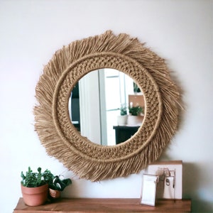 May include: A round mirror with a natural fiber frame and fringe. The frame is a light brown color and the fringe is a darker brown. The mirror is reflecting a room with a window and plants.
