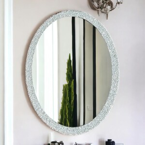 Puede incluir: Un espejo ovalado plateado con un marco texturizado brillante. El espejo est&aacute; colgado en una pared blanca sobre un lavabo de ba&ntilde;o.