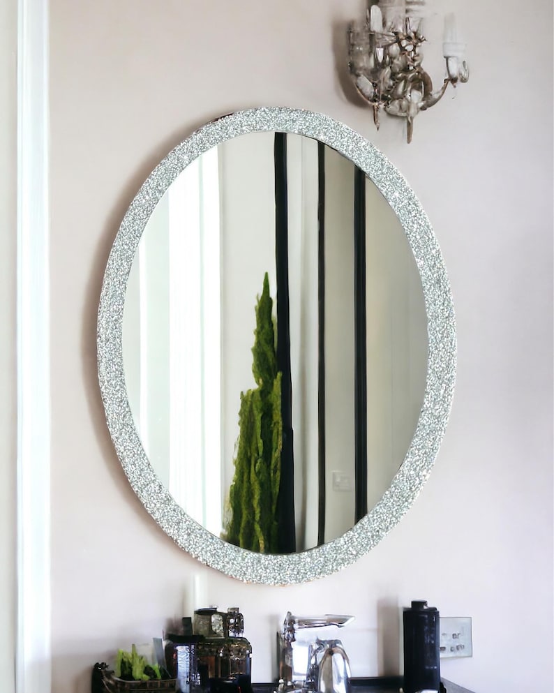 Puede incluir: Un espejo ovalado plateado con un marco texturizado brillante. El espejo est&aacute; colgado en una pared blanca sobre un lavabo de ba&ntilde;o.