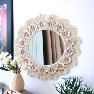 May include: A round mirror with a macrame frame and wooden beads. The frame is cream colored and has a fringe around the edge. The mirror is hanging on a white wall.