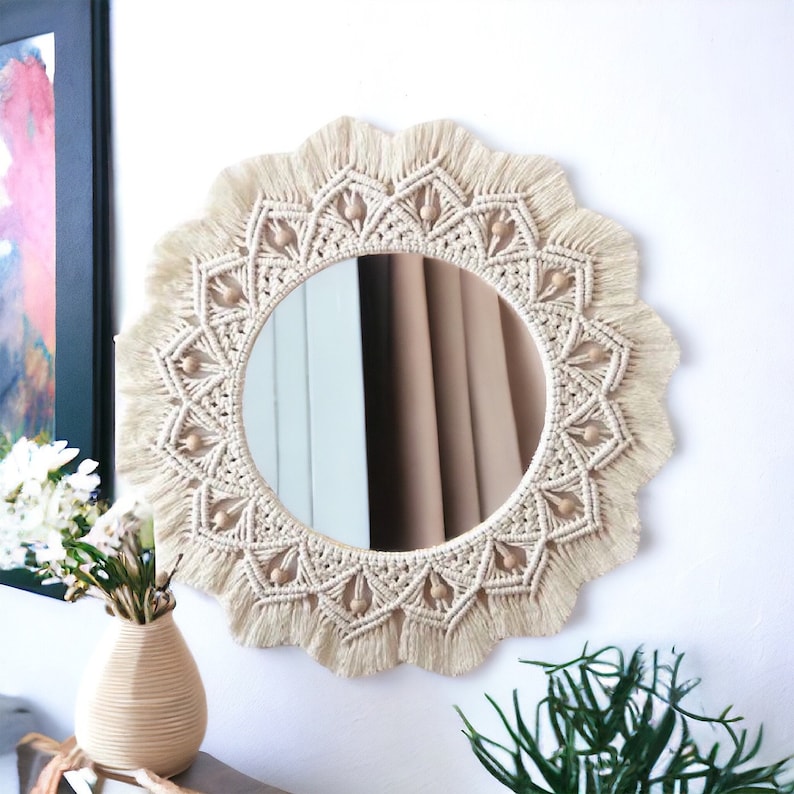 May include: A round mirror with a macrame frame and wooden beads. The frame is cream colored and has a fringe around the edge. The mirror is hanging on a white wall.