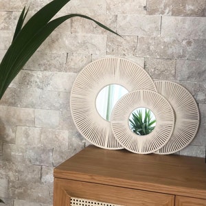 May include: Two round mirrors with a woven rope design, the larger mirror is behind the smaller mirror. The mirrors are cream colored and have a natural wood frame.