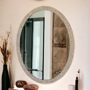 Puede incluir: Un gran espejo ovalado con un marco brillante de color plateado. El espejo est&aacute; colgado en una pared blanca sobre un estante con dos objetos decorativos.