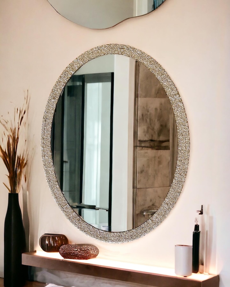 Puede incluir: Un gran espejo ovalado con un marco brillante de color plateado. El espejo est&aacute; colgado en una pared blanca sobre un estante con dos objetos decorativos.