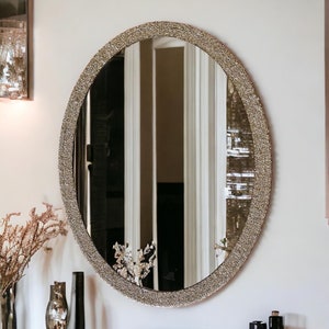 Puede incluir: Un espejo grande de forma ovalada con un marco decorativo dorado brillante. El espejo est&aacute; colgado en una pared blanca.
