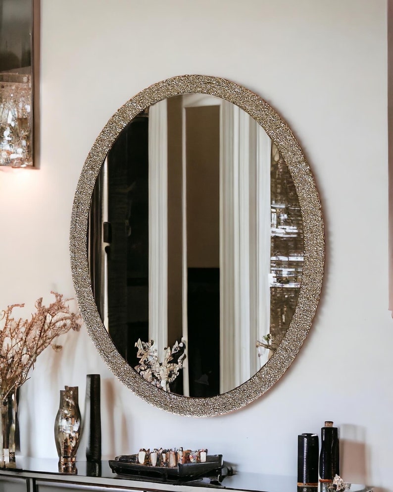 Puede incluir: Un espejo grande de forma ovalada con un marco decorativo dorado brillante. El espejo est&aacute; colgado en una pared blanca.