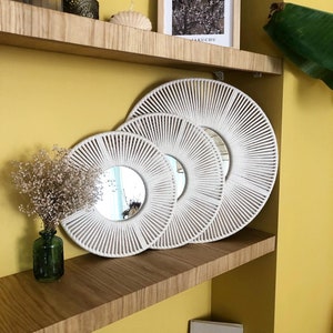 May include: Three white macrame wall mirrors of varying sizes are displayed on a wooden shelf against a yellow wall. The mirrors are round and have a sunburst design.