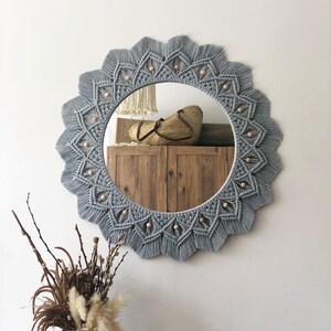 May include: A gray macrame mirror with a round wooden frame and a beaded border. The mirror is hanging on a white wall.