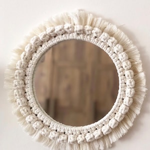 May include: A round mirror with a white macrame frame and fringe. The mirror is surrounded by a thick rope border with a knotted design.