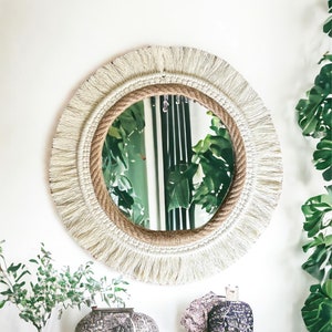 May include: A round mirror with a white fringe border and a natural rope frame. The mirror is reflecting a green plant.