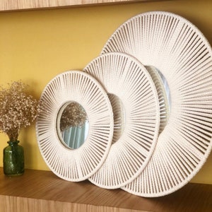 May include: Three round mirrors with a sunburst design made of woven rope. The mirrors are nested inside each other, with the largest mirror in the back and the smallest mirror in the front. The mirrors are a cream color and have a natural wood shelf behind them.