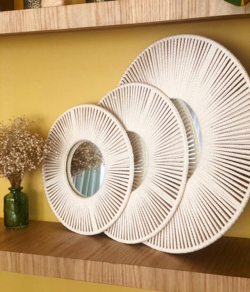 May include: Three round mirrors with a sunburst design made of woven rope. The mirrors are nested inside each other, with the largest mirror in the back and the smallest mirror in the front. The mirrors are a cream color and have a natural wood shelf behind them.