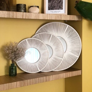 May include: Three white rope mirrors with a sunburst design, arranged in a decreasing size order on a wooden shelf. The mirrors are on a shelf with a yellow wall in the background.