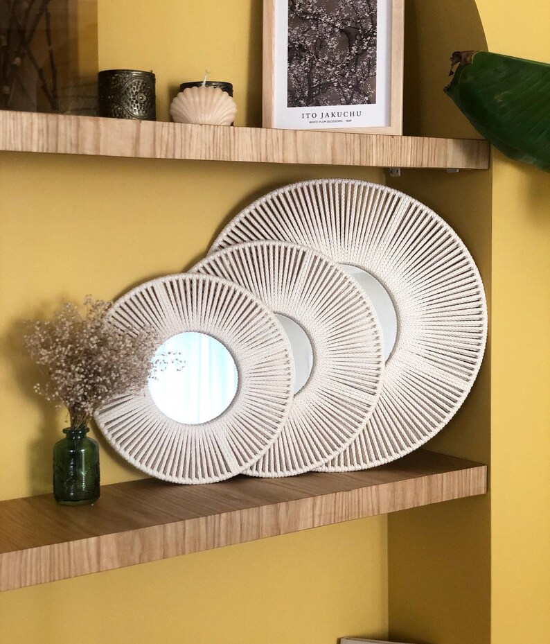 May include: Three white rope mirrors with a sunburst design, arranged in a decreasing size order on a wooden shelf. The mirrors are on a shelf with a yellow wall in the background.
