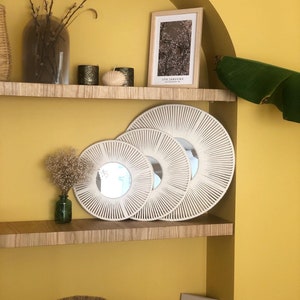 May include: Three round macrame mirrors of different sizes, with a white rope design, are displayed on a wooden shelf against a yellow wall. A framed print with the text "ITO JAKUCHU" is on the shelf above the mirrors.