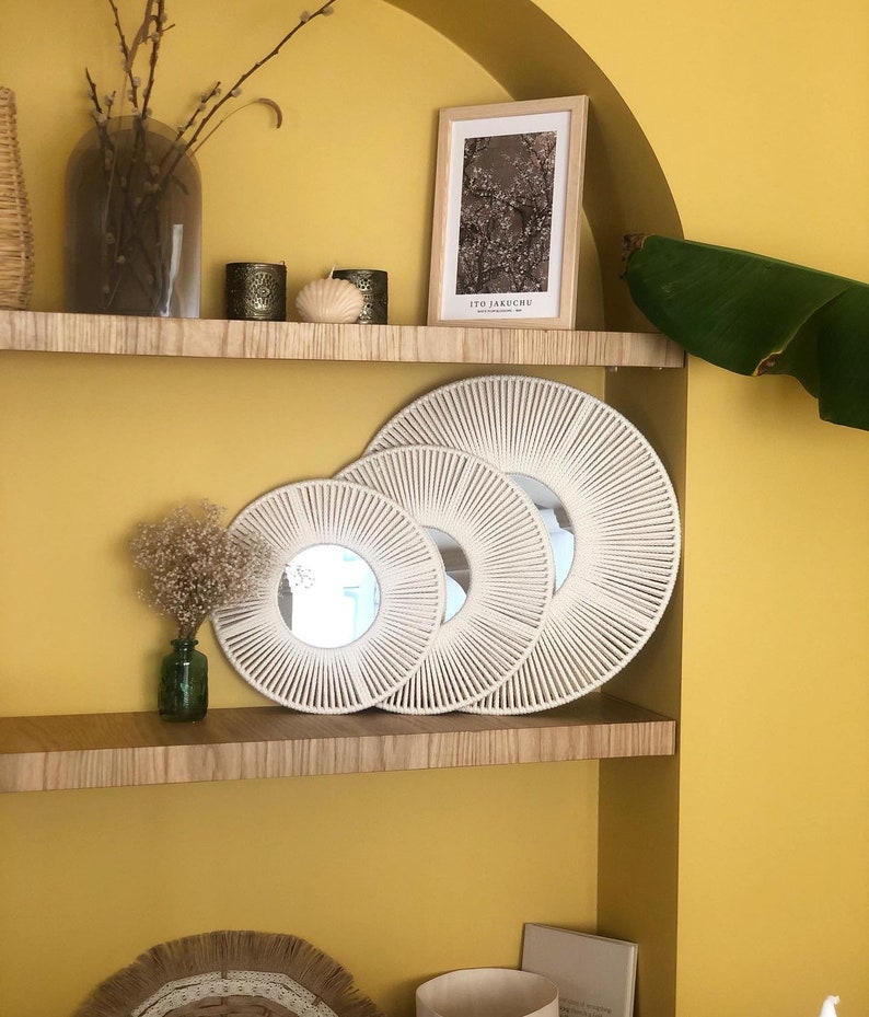 May include: Three round macrame mirrors of different sizes, with a white rope design, are displayed on a wooden shelf against a yellow wall. A framed print with the text "ITO JAKUCHU" is on the shelf above the mirrors.