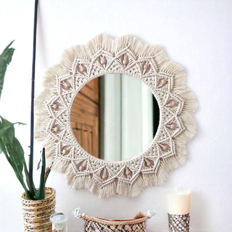 May include: A round macrame mirror with a white cotton cord and wooden beads. The mirror has a fringe border and a mandala design.
