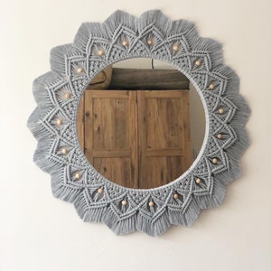 May include: A gray macrame mirror with a circular wooden frame and a beaded design. The mirror is hanging on a white wall.