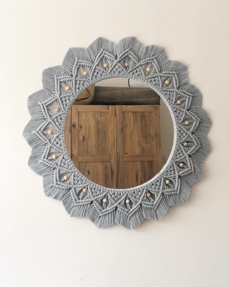 May include: A gray macrame mirror with a circular wooden frame and a beaded design. The mirror is hanging on a white wall.