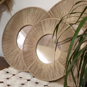 May include: Three round mirrors with a natural rope frame. The mirrors are nested inside each other, with the largest mirror in the back and the smallest mirror in the front. The mirrors are on a white lace tablecloth.