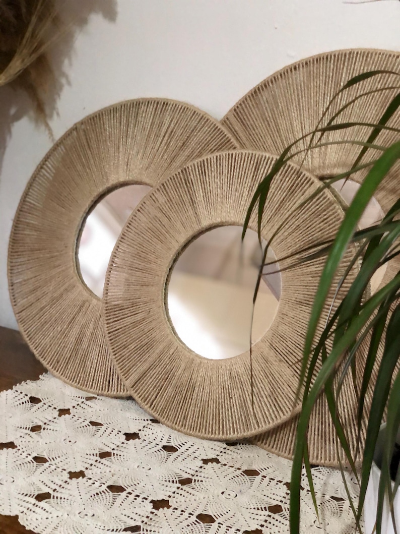 May include: Three round mirrors with a natural rope frame. The mirrors are nested inside each other, with the largest mirror in the back and the smallest mirror in the front. The mirrors are on a white lace tablecloth.